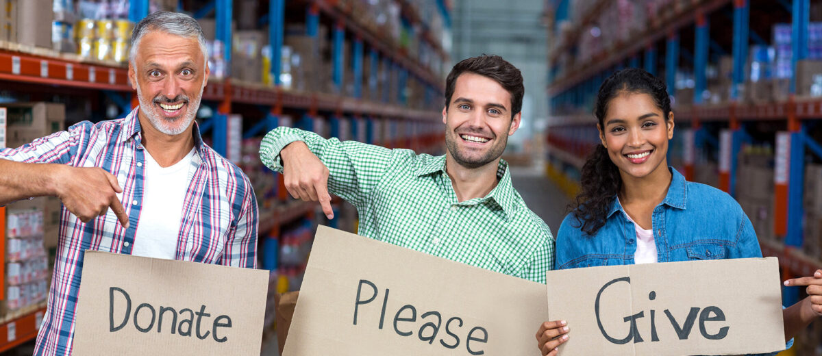 photo of three smiling people standing in a warehouse holding signs that read donate, please, and give